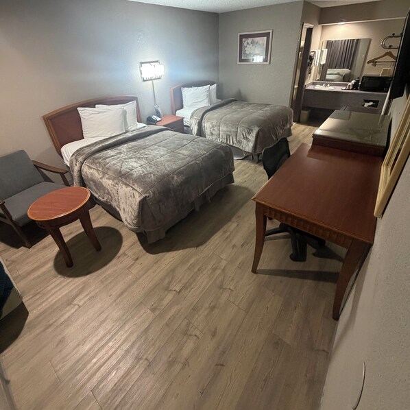 A hotel room with two beds, a small round table, a wooden desk, a mirror, and a bathroom with a sink and mirror.