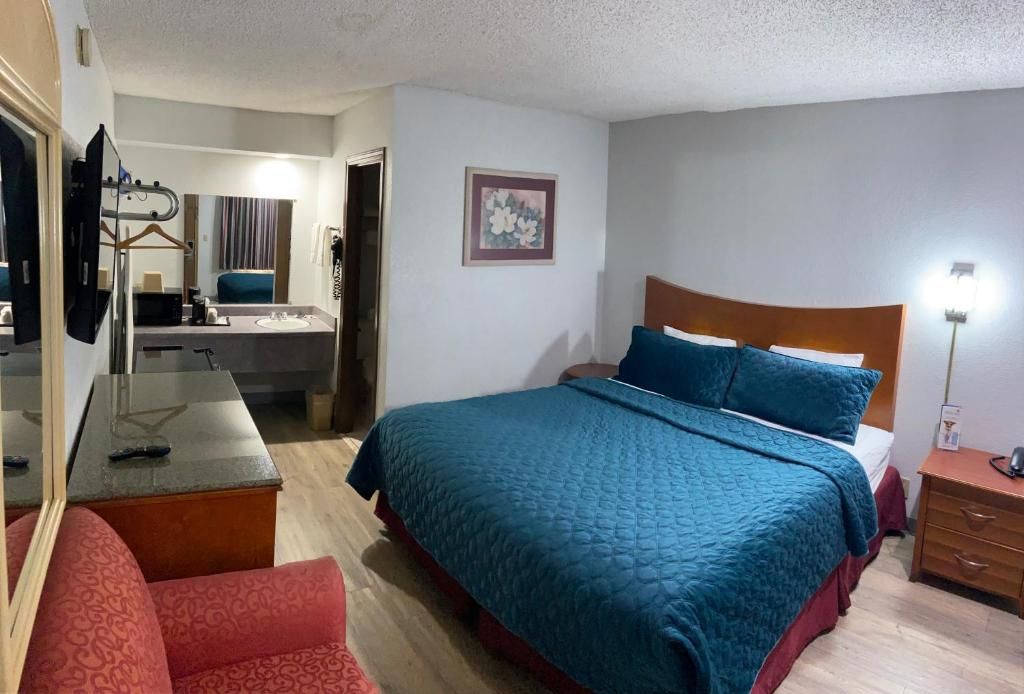 A hotel room with a bed, a desk with a chair, a mirror, a bedside table, and a wall-mounted TV.