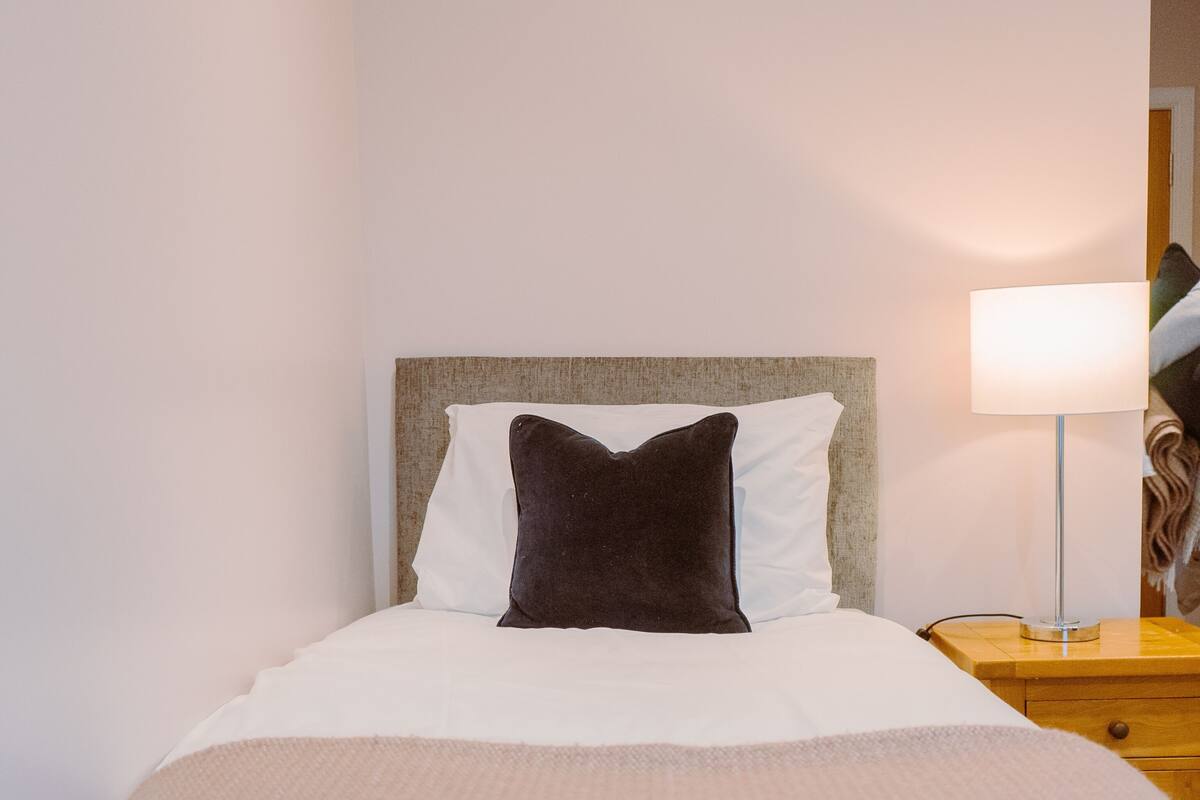 A neatly made bed with a white duvet and a dark brown pillow, a bedside lamp, and a wooden nightstand.