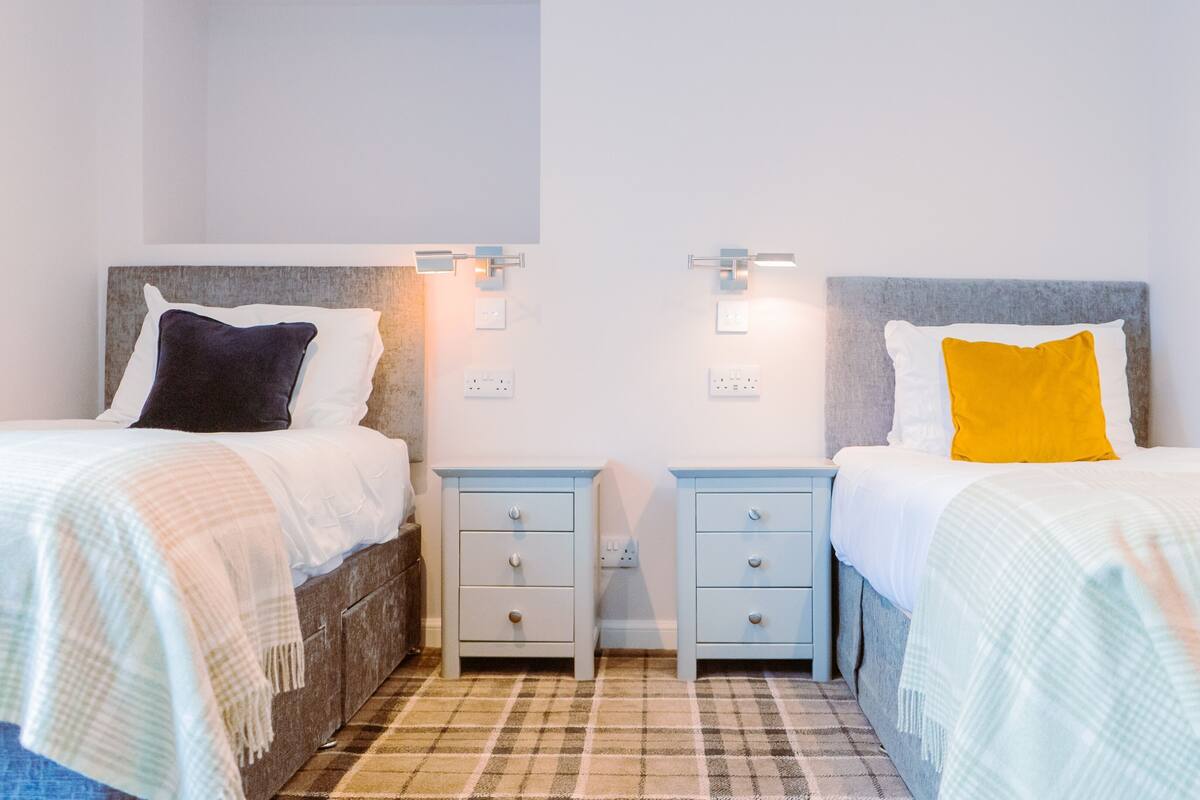 A room with two beds, each with a pillow and a bedside table. There are wall sockets and a light switch on the wall.