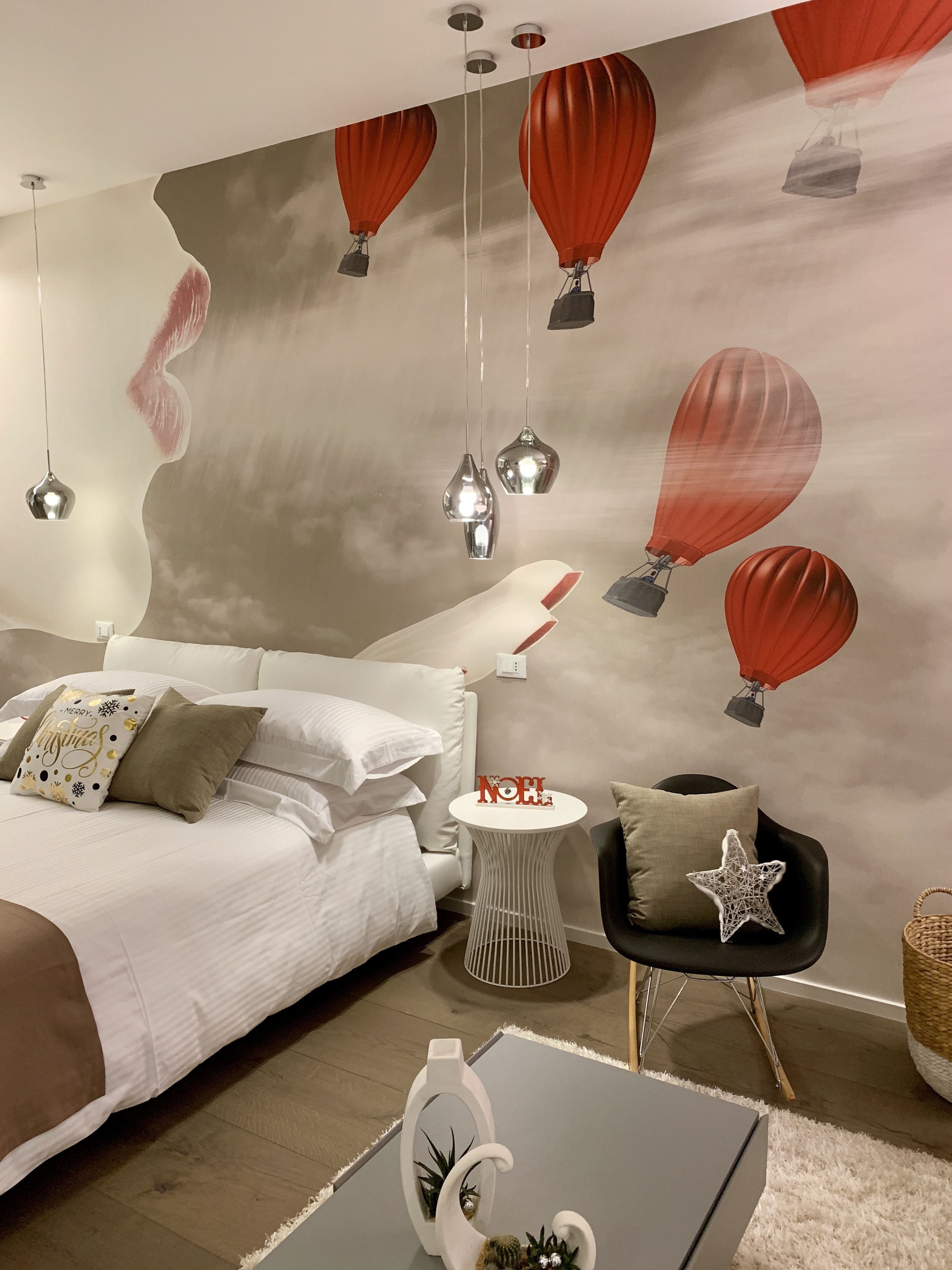 A modern bedroom with a bed, bedside tables, a small side table with a 'NOEL' decoration, and a wall mural of hot air balloons.