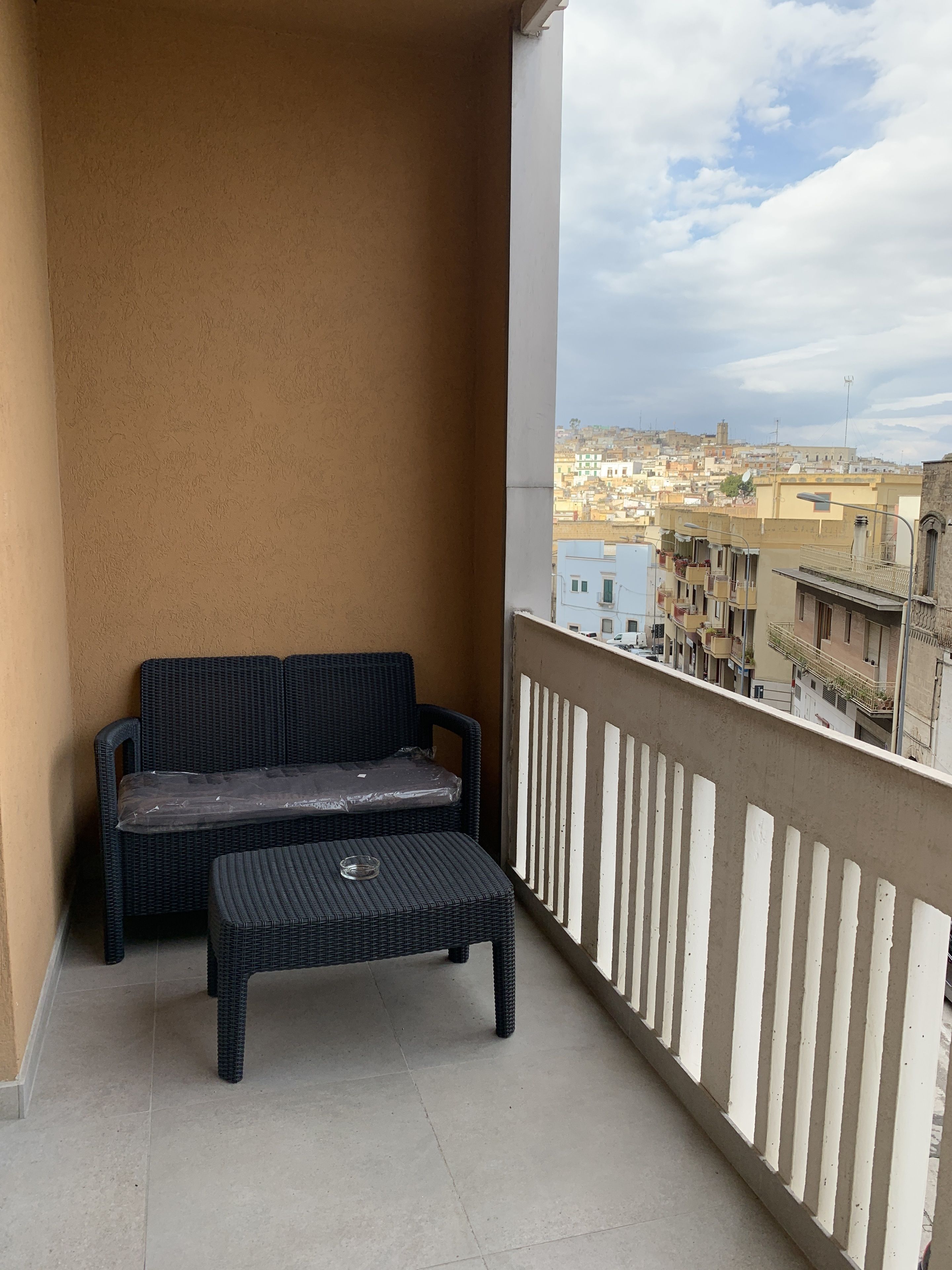 A balcony with a wicker sofa and a small table, overlooking a cityscape with buildings and a clear sky.