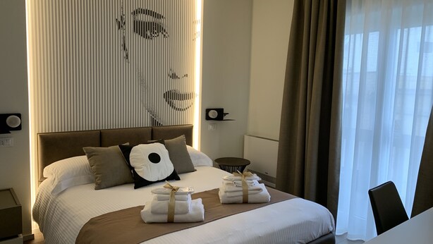 A hotel room with a large bed, a bedside table, a wall-mounted speaker, and a window with curtains. - Agorà B&B Urban Relais (Canosa di Puglia)