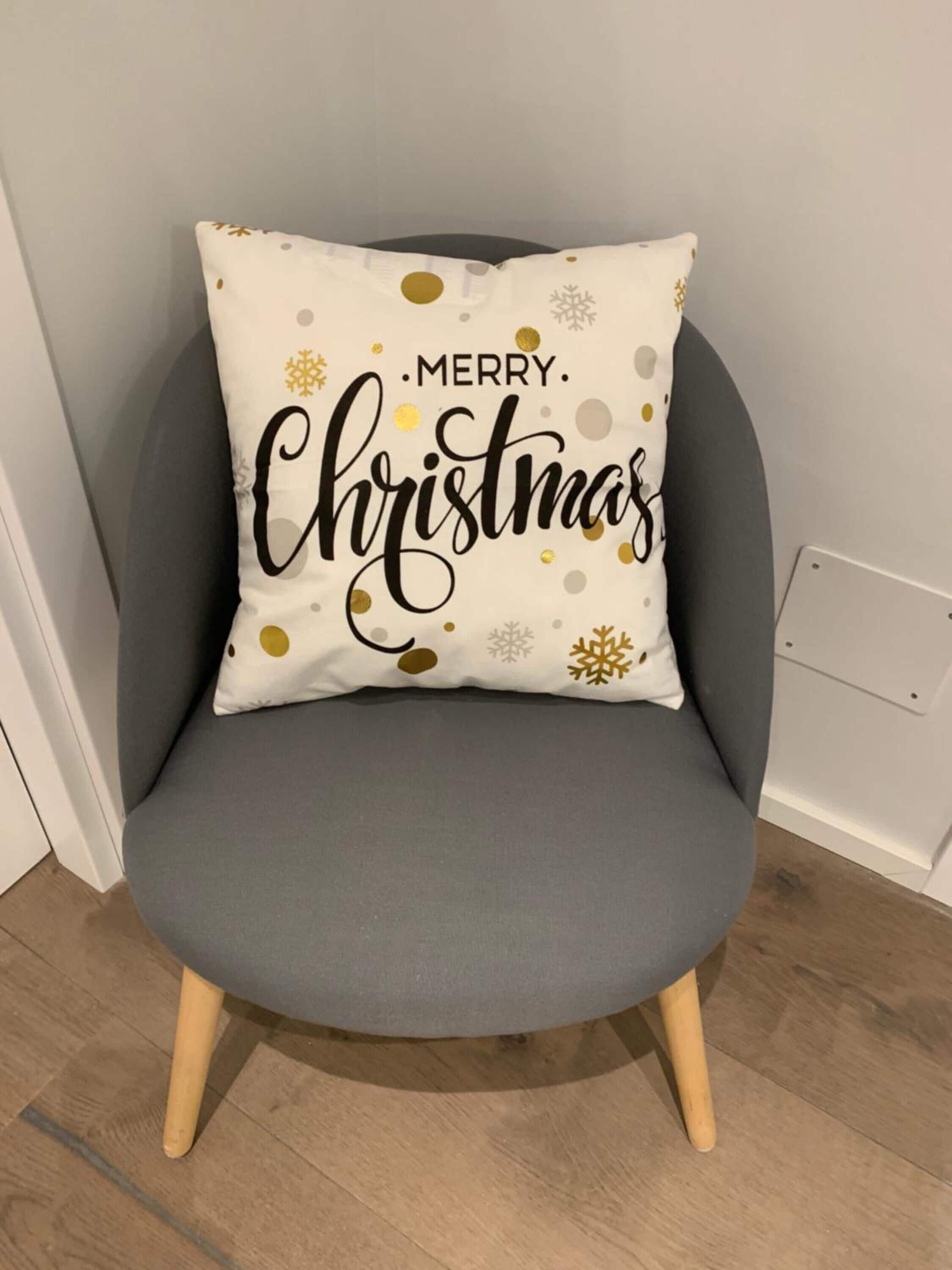 A grey armchair with a white cushion featuring 'Merry Christmas' text and decorative elements.