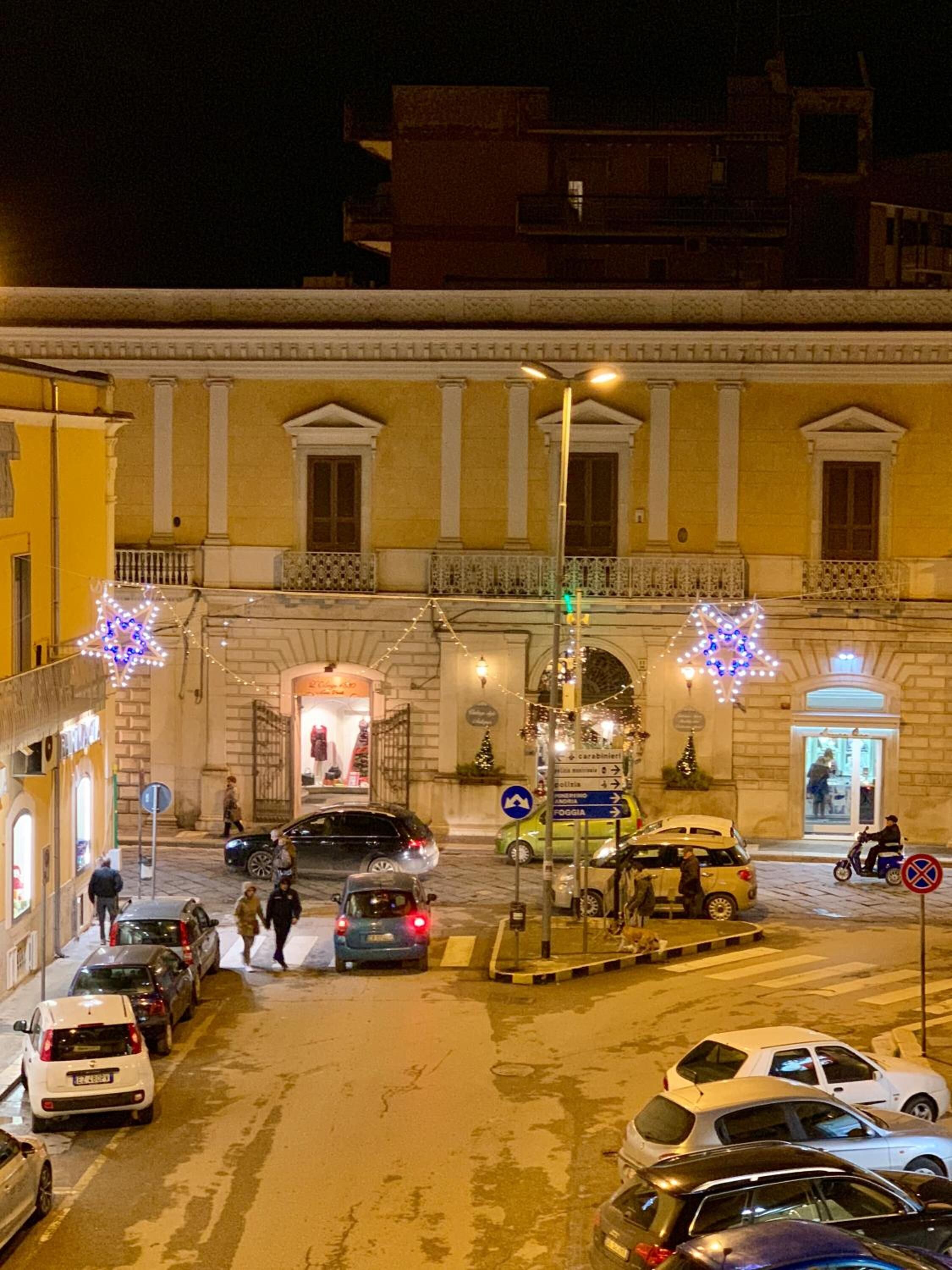 A street view at night with parked cars, a pedestrian crossing, and a building with classical architecture.