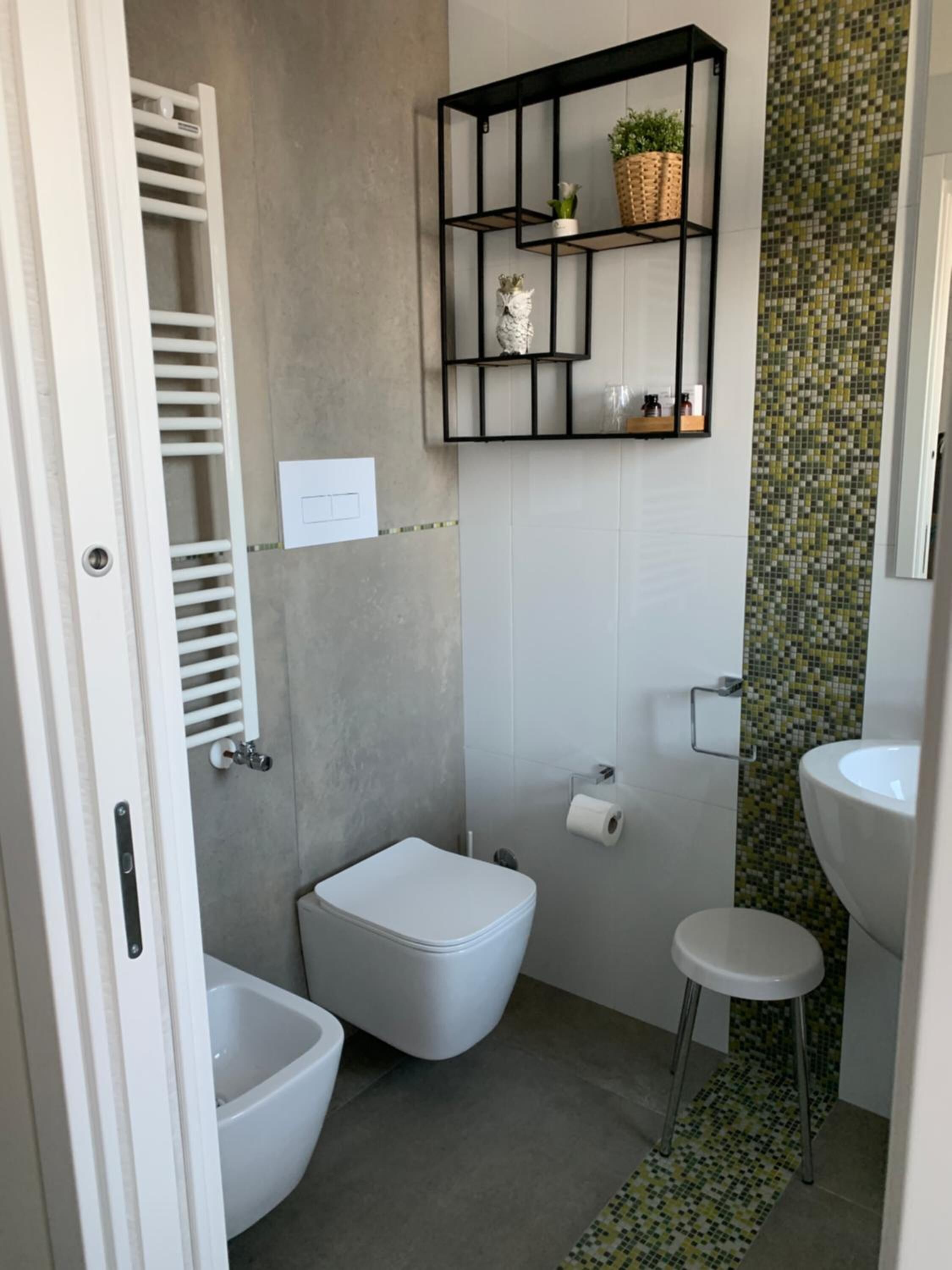 Et lille badeværelse med toilet, bidet og en lille skammel. Der er en hylde med planter og en kurv over toilettet.