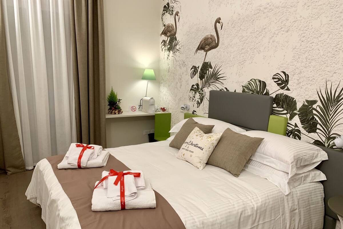 Quarto de hotel com uma cama bem arrumada, mesa de cabeceira com abajur, parede com mural tropical e planta verde na mesa de cabeceira.