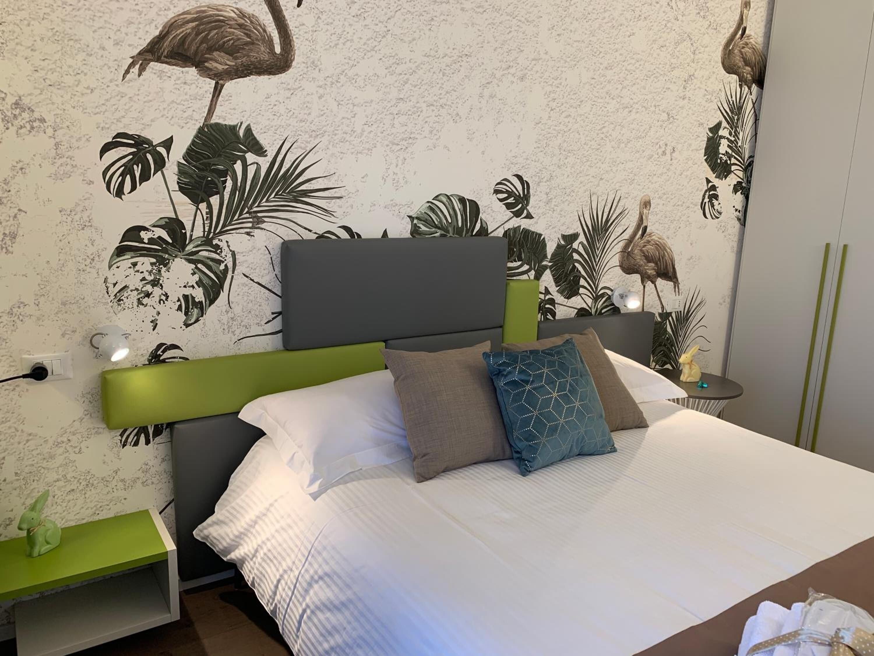 A bedroom with a bed, green nightstands, a wall with tropical leaf and bird decals, and a small table with a lamp.