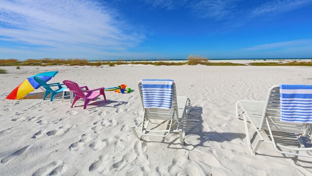 A beach with two white lounge chairs with blue towels, colorful beach chairs, and a beach umbrella. - Cozy cottage in Gulf Gate to relax year-round in beautiful Sarasota. (Sarasota)