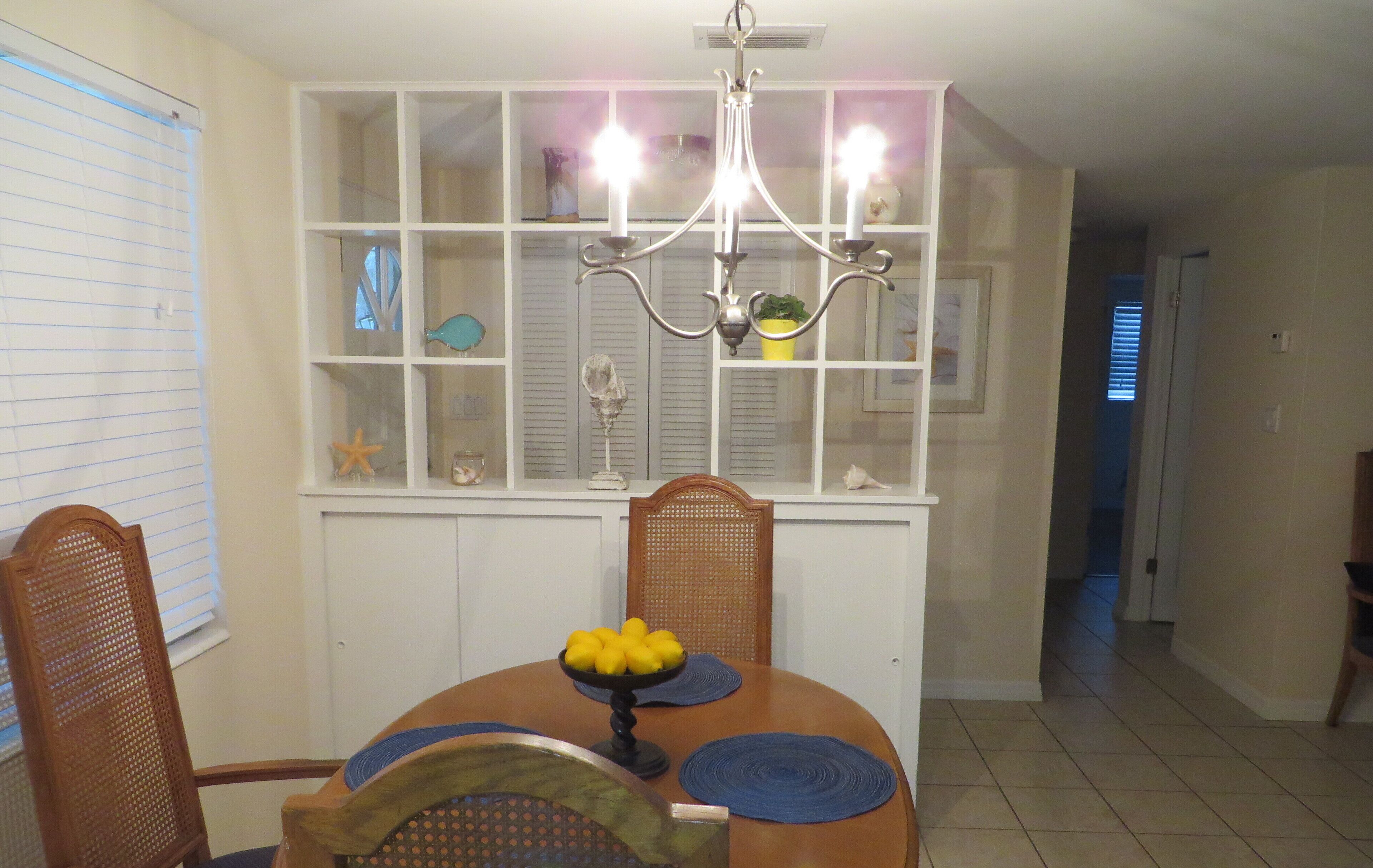 Un comedor con una mesa de madera, sillas, una araña y una estantería decorativa con varios objetos.