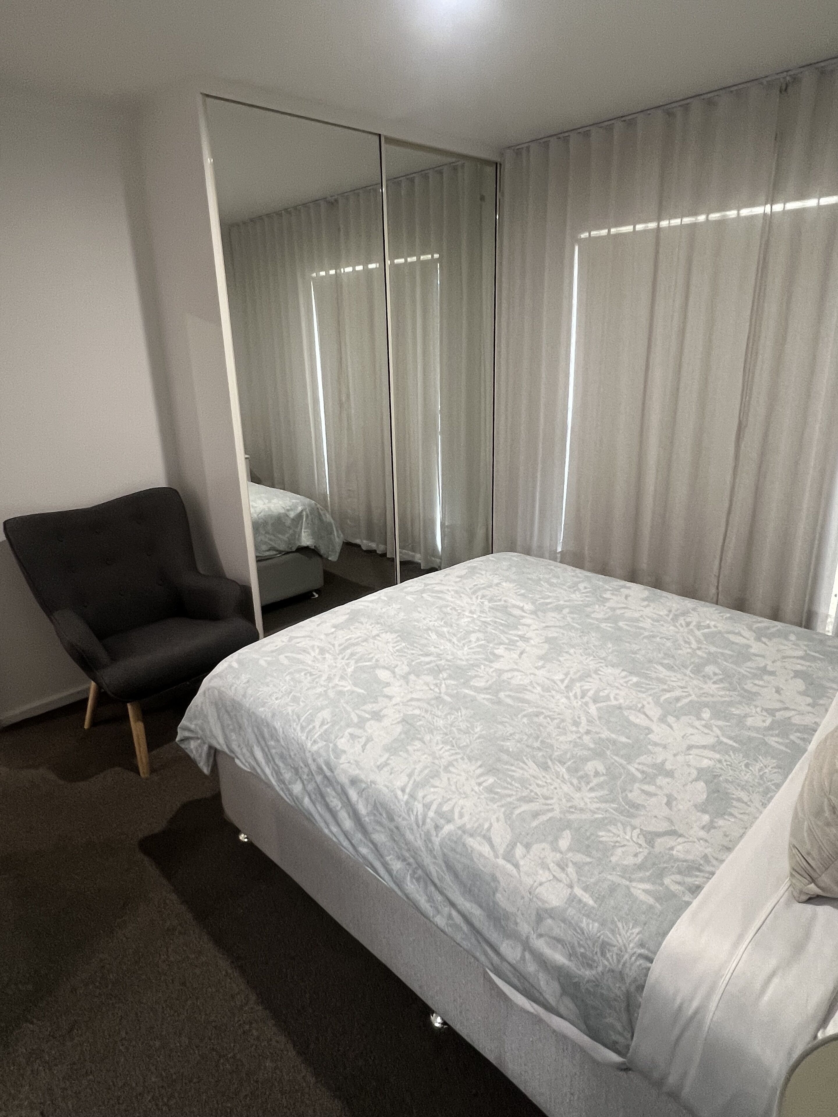 A bedroom with a bed, a chair, a large mirror, and a window with curtains.