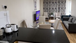 A compact kitchen with a dishwasher, a small dining area with a glass table, and a living room with a sofa and a TV. - ADELAIDE CENTRAL APARTMENT - 3BR, 2BATH & CARPARK (Adelaide)