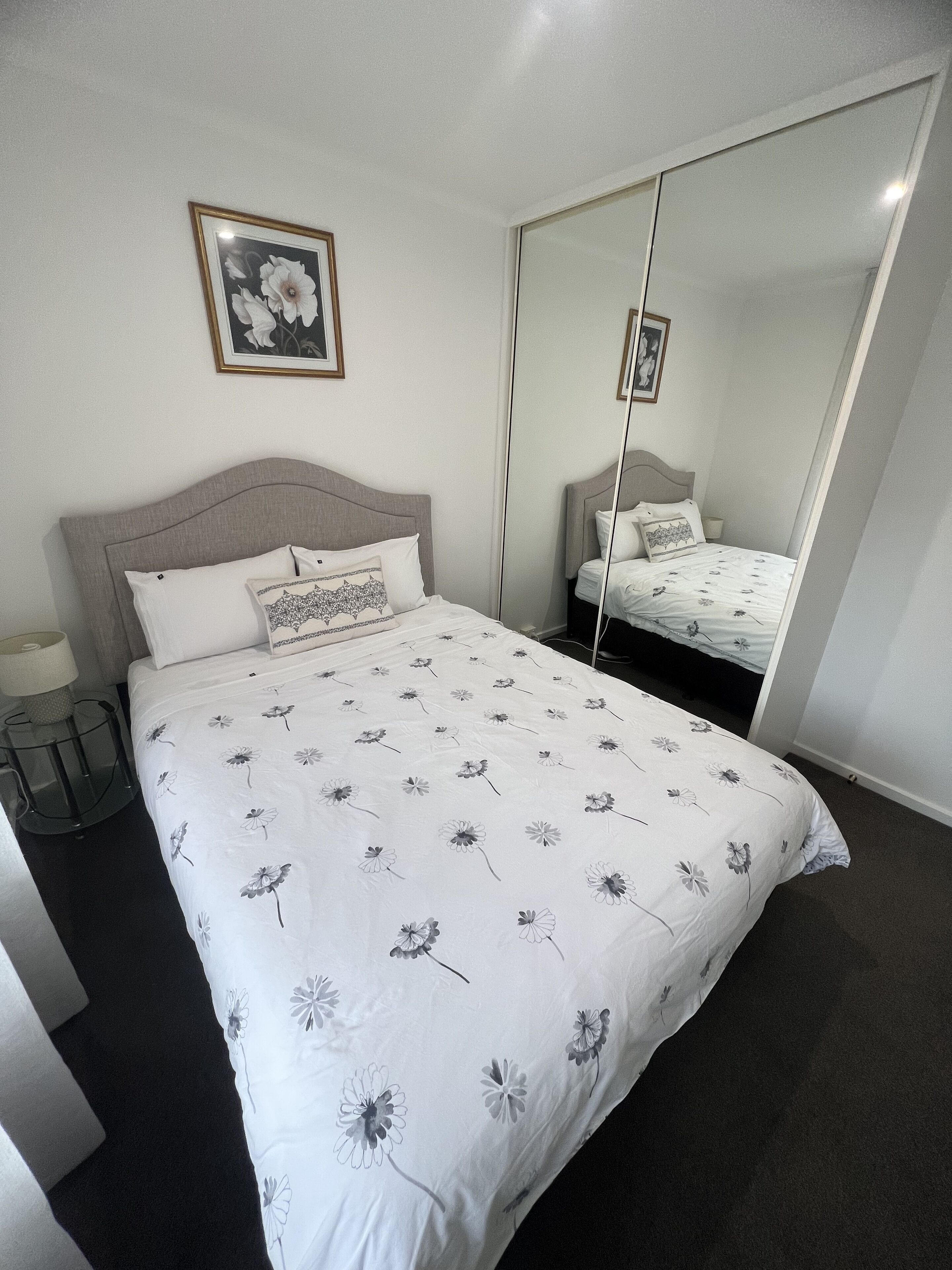 A neatly made bed with a floral bedspread, a bedside lamp, and a framed picture on the wall.
