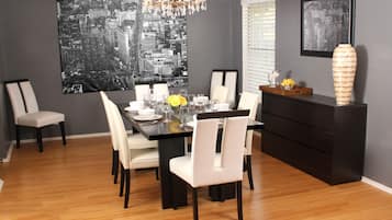 Un coin repas avec une table noire, des chaises blanches, un lustre et un tableau représentant un paysage urbain accroché au mur.