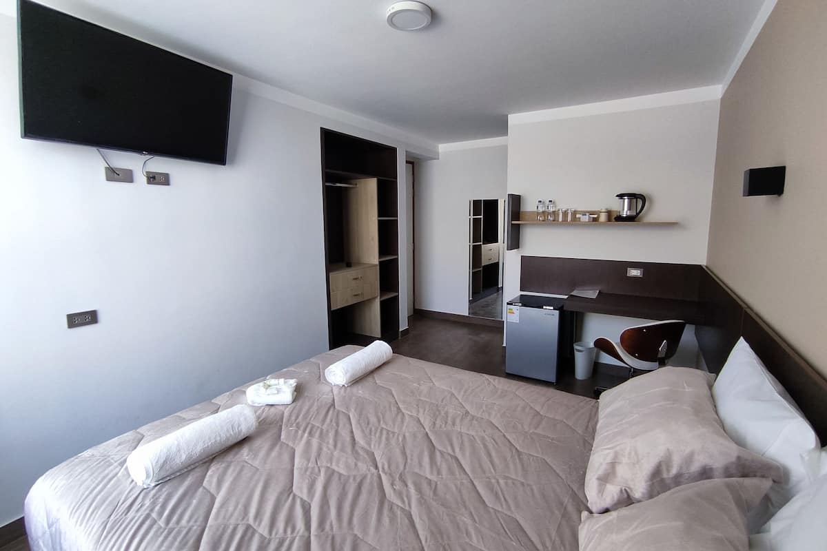A hotel room with a bed, a desk, a chair, a flat-screen TV mounted on the wall, and a small kitchenette area.