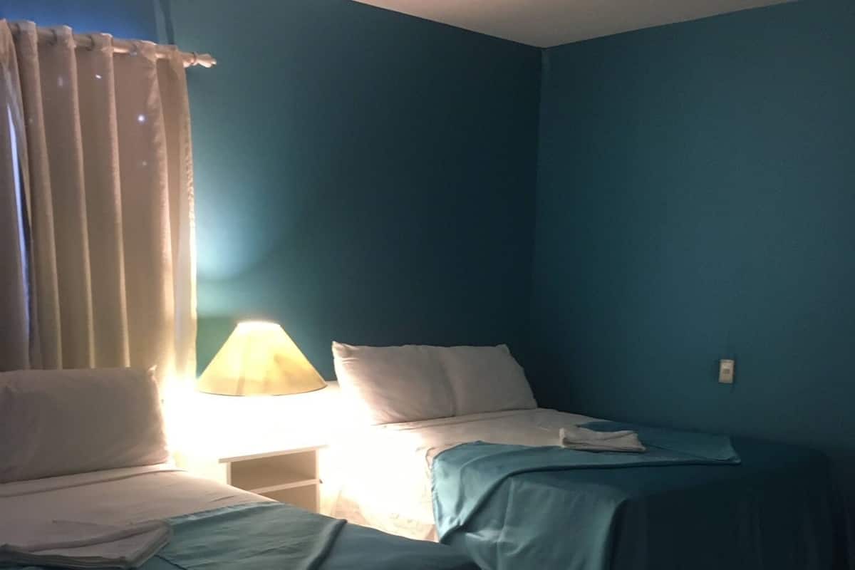 Una habitación con dos camas, cada una con cubrecama azul y almohadas blancas. Hay una mesita de noche con una lámpara entre las camas.