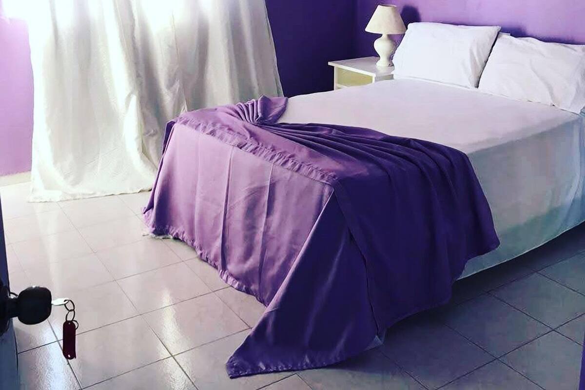 Un dormitorio con cama, cortinas blancas, una mesita de noche con lámpara y un llavero en el suelo.