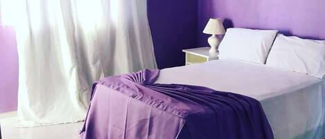 A bedroom with a bed, white curtains, a nightstand with a lamp, and a keychain on the floor.