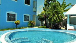 Una piscina con borde de azulejos azules, rodeada por una terraza blanca. Se observan palmeras y un edificio con paredes azules y ventanas blancas.