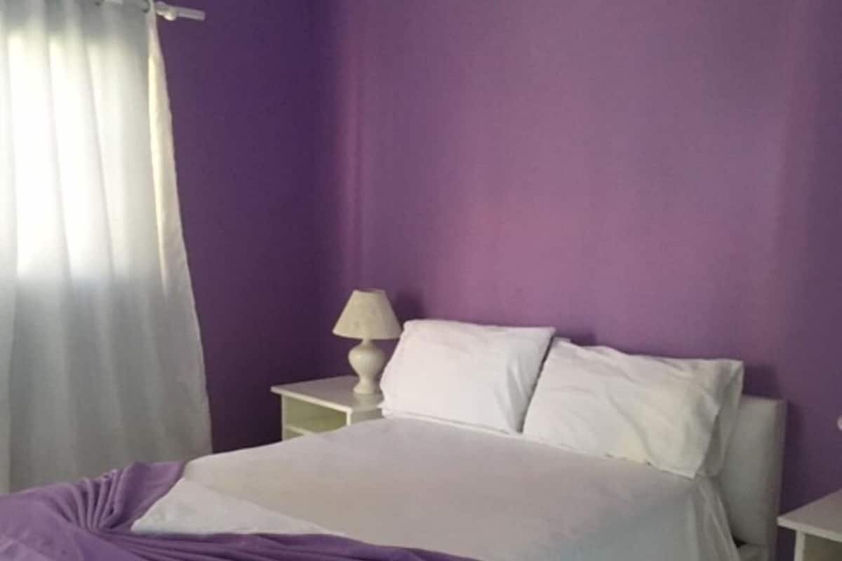 Un dormitorio con cama, cortinas blancas, una mesita de noche con lámpara y una pared morada.