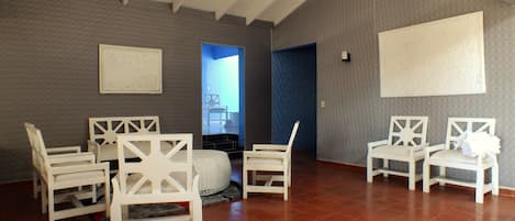A spacious room with a high ceiling and red-tiled floor, featuring a seating area with white chairs and a small round table.