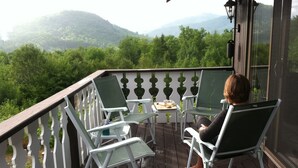 A balcony with two green chairs and a small table, overlooking a forested area with mountains in the distance. - Stunning views of Mt Washington and the presidentials. (Bartlett)
