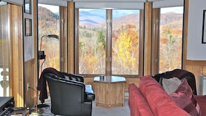 A room with a large window offering a view of autumn foliage and mountains. There is a black leather chair, a red sofa, and a small wooden table. - Stunning views of Mt Washington and the presidentials. (Bartlett)