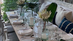 A long dining table set with plates, glasses, and cutlery, with a view of the beach and palm trees through large windows. - Casa Violeta (Tulum)