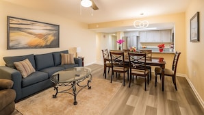 A living room with a blue sofa, a glass coffee table, and a dining area with wooden chairs and a table. - Beautiful Direct Oceanfront 2-BR Condo on Top Floor at Broadwalk, Downtown (Myrtle Beach)