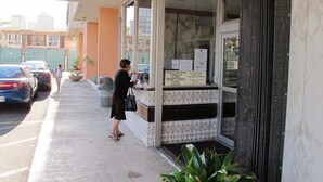 Une femme se tient derrière un comptoir d’accueil, avec un panneau indiquant la présence d’une caméra de surveillance, un pot de fleurs et des voitures garées devant un bâtiment dont l’entrée est recouverte de carreaux.