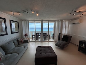 A spacious living room with a grey sofa, a dark ottoman, and a view of the ocean through large sliding glass doors. - BEACHFRONT ON BEACH - OCEAN VIEW BALCONY WITH POOL & CASINO!  DISCOUNT (Condado)