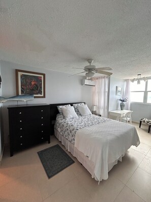 A bedroom with a bed, a dresser, a ceiling fan, an air conditioning unit, a desk, a chair, a window with curtains, and a painting on the wall.