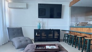Un salon avec un canapé gris, une table basse sur laquelle se trouve un plateau, un téléviseur fixé au mur, un meuble noir et un coin bar avec des tabourets.