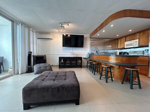 A modern living room with a large grey ottoman, a flat-screen TV mounted on the wall, and a kitchen area with a bar counter and stools.