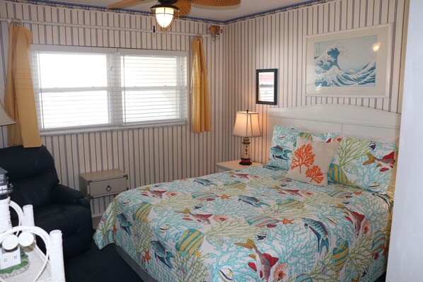 A bedroom with striped wallpaper, a bed with a colorful fish pattern, a black chair, a nightstand with a lamp, and a window with curtains.