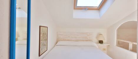 A bedroom with a skylight, a bed with a white bedspread, a bedside table with a lamp, and a framed picture on the wall.