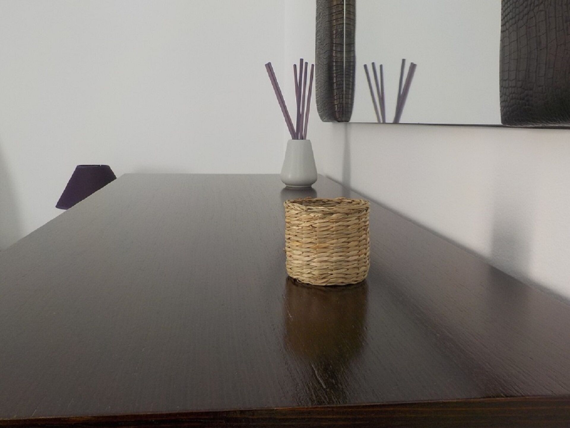 A dark wooden table with a woven basket and a white vase with reed sticks.