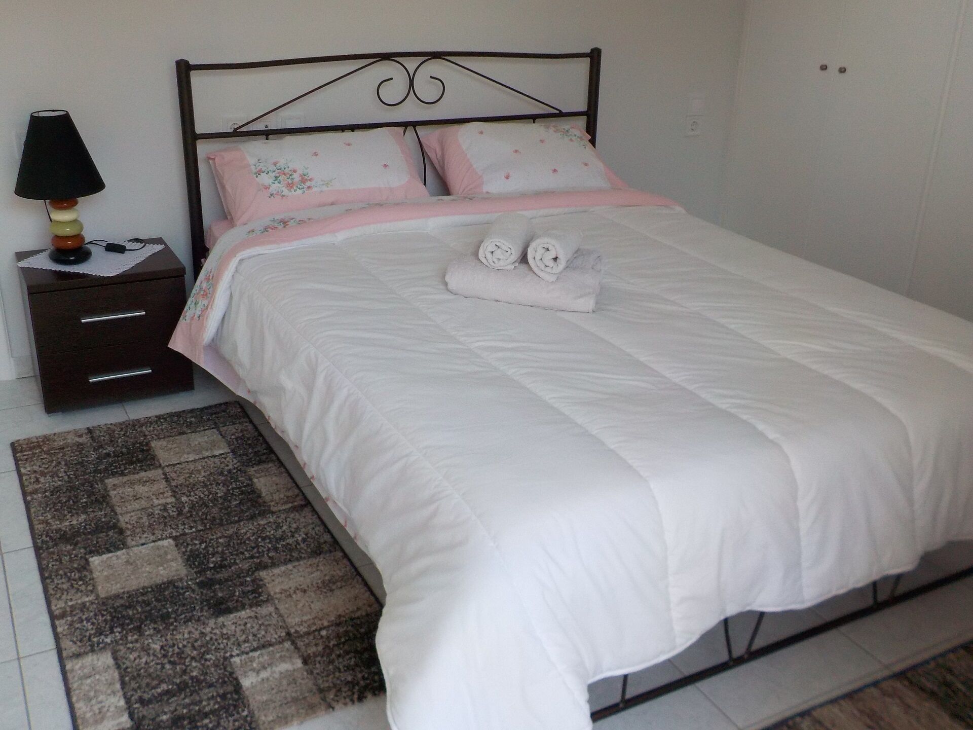 A neatly made bed with white bedding and pink floral pillows, a dark wooden nightstand with a lamp, and a patterned rug on a tiled floor.