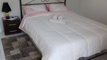 A neatly made bed with white bedding and pink floral pillows, a dark wooden nightstand with a lamp, and a patterned rug on a tiled floor.