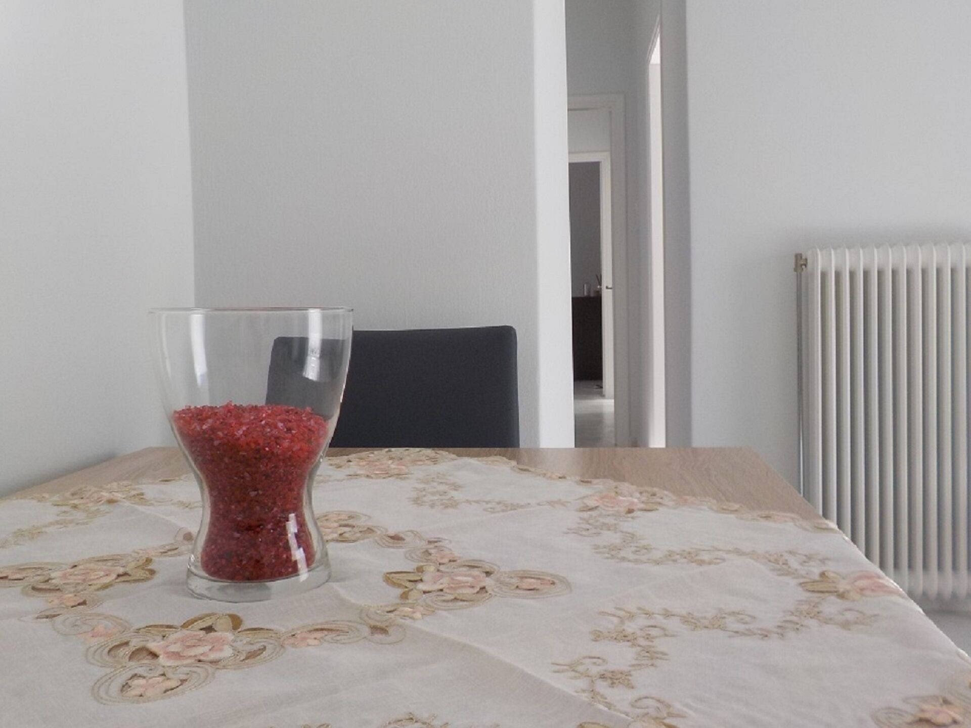 Uma mesa de jantar com toalha florida, um copo com grânulos vermelhos e uma cadeira.