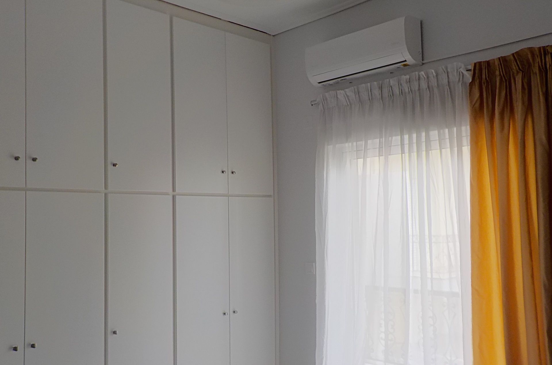 Een kamer met een witte kledingkast, een airconditioning en een raam met lichte witte gordijnen en een gele stofsluier.
