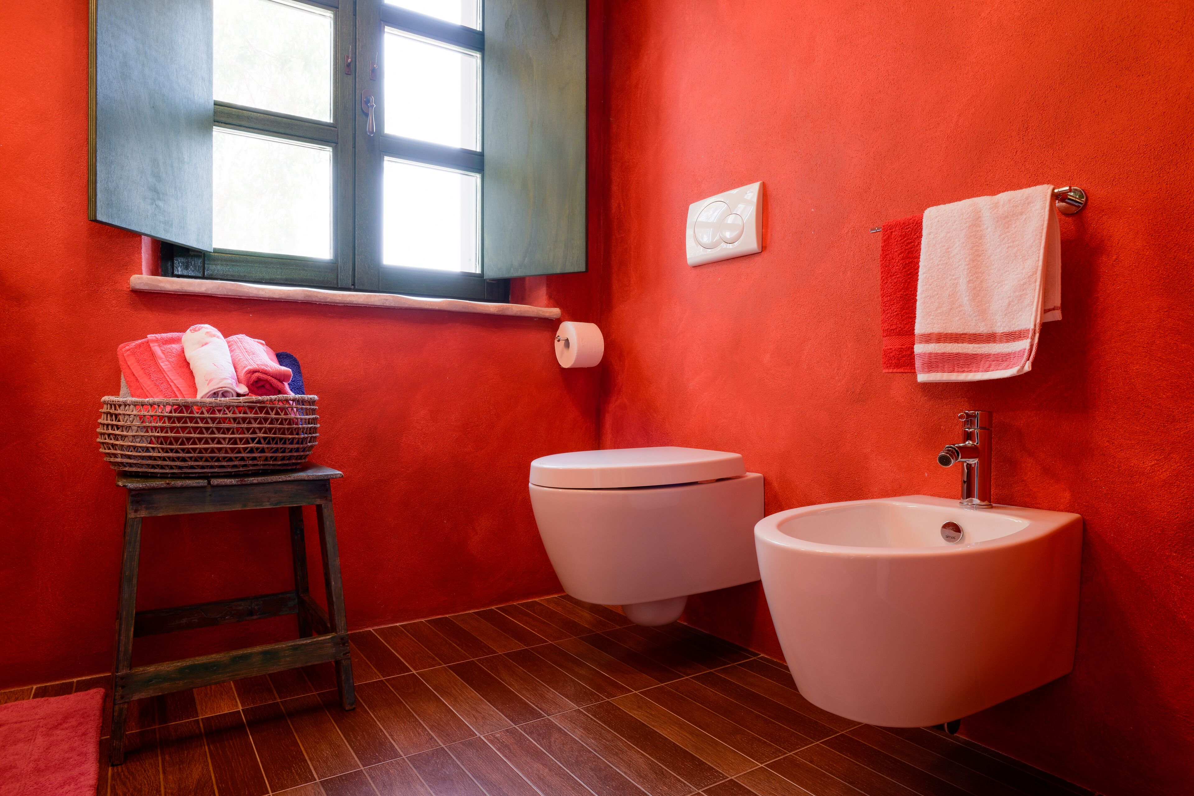 Ein Badezimmer mit roter Wand, einem Fenster, einer Toilette, einem Bidet, einem Waschbecken und einem Handtuchhalter.