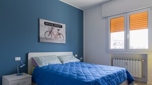 A bedroom with a blue bedspread, a nightstand with a lamp, a wall-mounted bicycle sign, and a window with blinds.