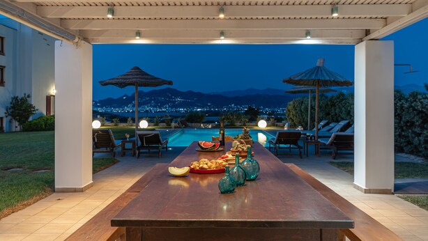 An outdoor dining area with a pool view, a wooden table set with food, and two umbrellas. - Aurora Villa with private pool & Jacuzzi - Naxos (1)