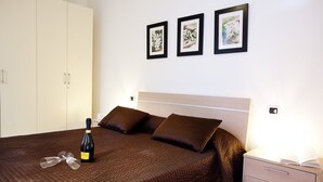 Ein ordentlich bezogenes Bett mit einer braunen Steude, einer Weinflasche und zwei Gläsern darauf. Über dem Bett hängen drei gerahmte Bilder an der Wand.