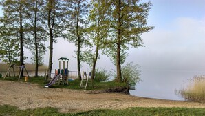 A playground with a slide and swings, a sandy area, and a body of water in the background. - Cottage in Masuria. Mazury famous for its 2,000 lakes. (Gizycko)