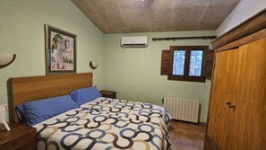 A bedroom with a bed, a radiator, an air conditioning unit, a window with shutters, and a door.