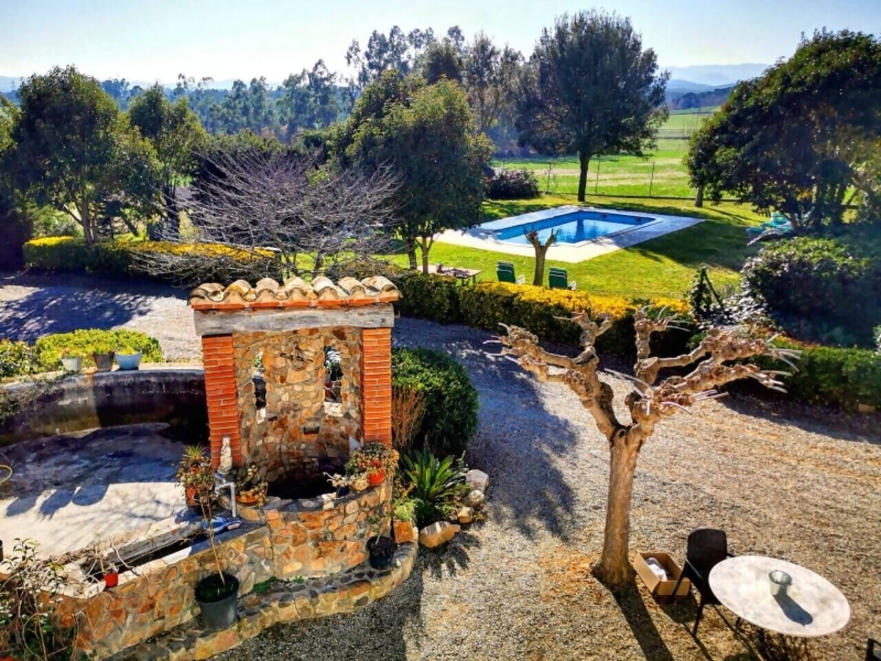 A stone structure with a small pool, surrounded by a landscaped garden with a tree and a clear view of a swimming pool in the background.