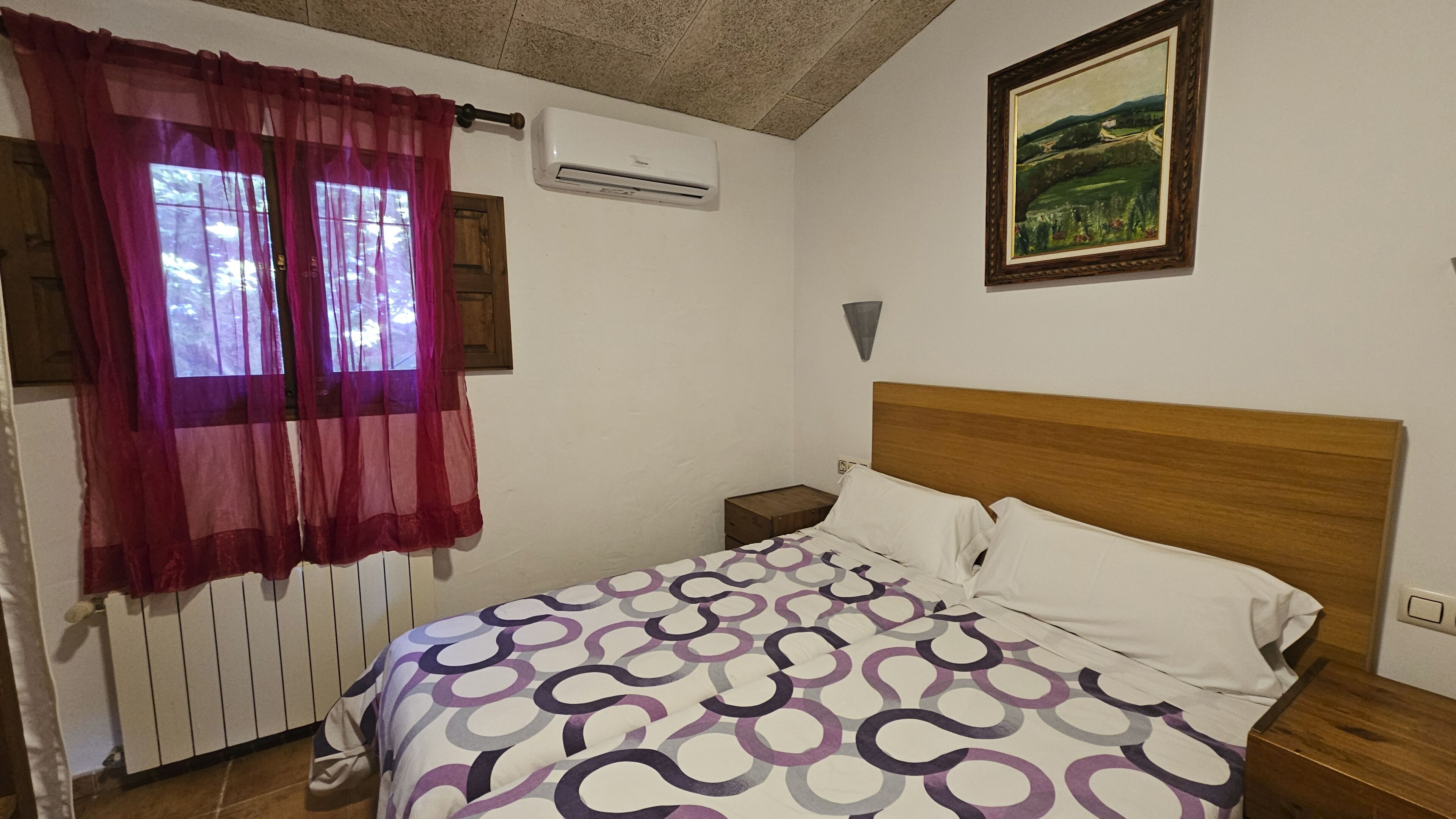 A bedroom with a bed, a window with sheer curtains, an air conditioning unit, a nightstand, and a painting on the wall.