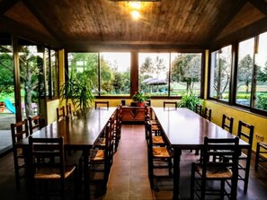 A long dining table with chairs, surrounded by large windows offering a view of greenery.